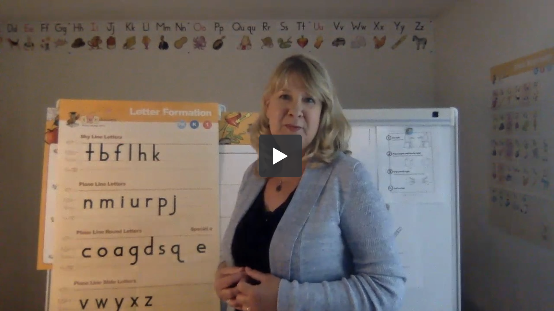 A woman is standing in a classroom with a large poster titled Letter Formation displaying letters. She wears a light blue blazer and smiles at the camera. The alphabet is displayed on the wall behind her. The play button in the center suggests it's a video.